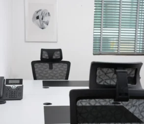 Empty office desk with chairs, phone, and window blinds in background.